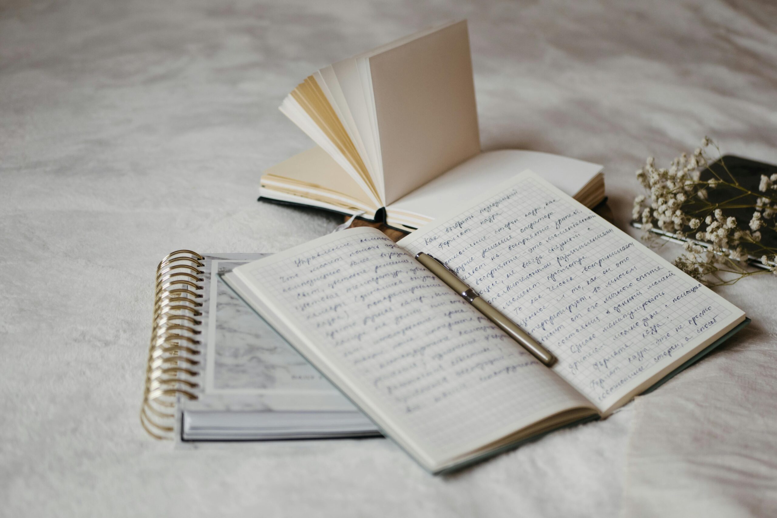 Close-up of open notebooks and a pen on a soft bed surface with delicate dried flowers.